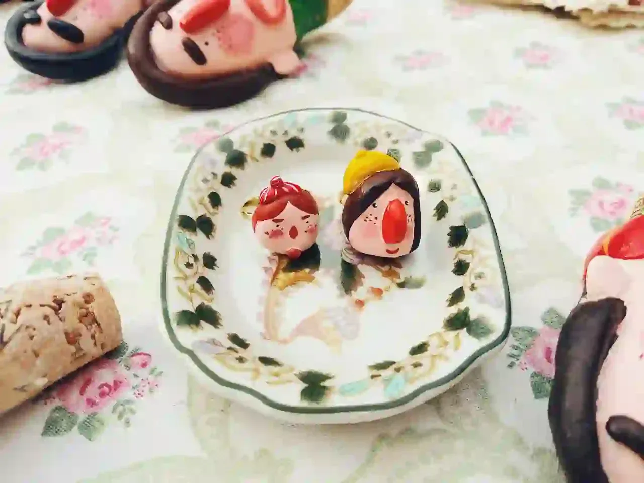 Small clay characters and a wine stopper arranged on a patterned plate