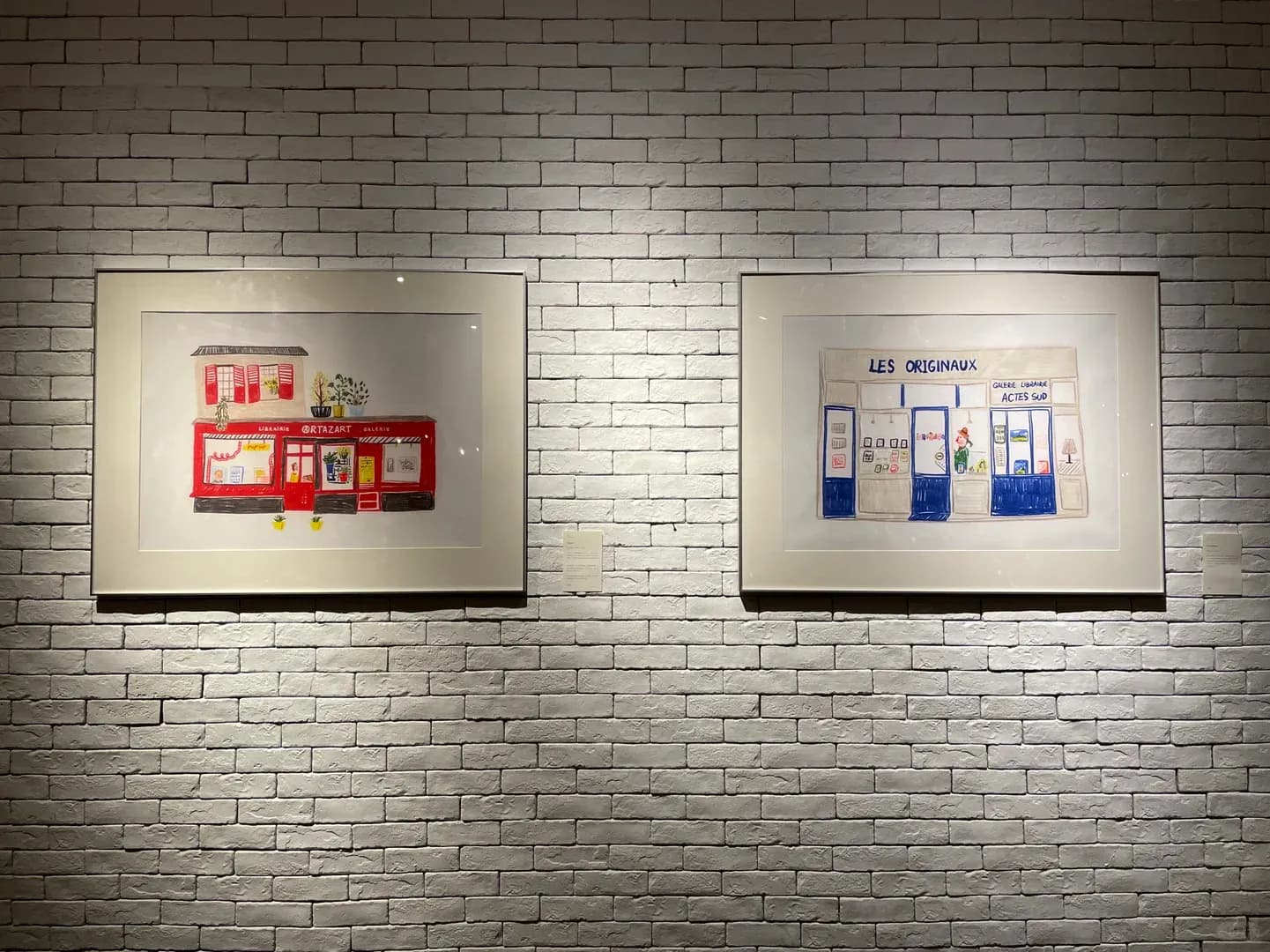 Two framed Paris bookshop illustrations shown on a white brick wall